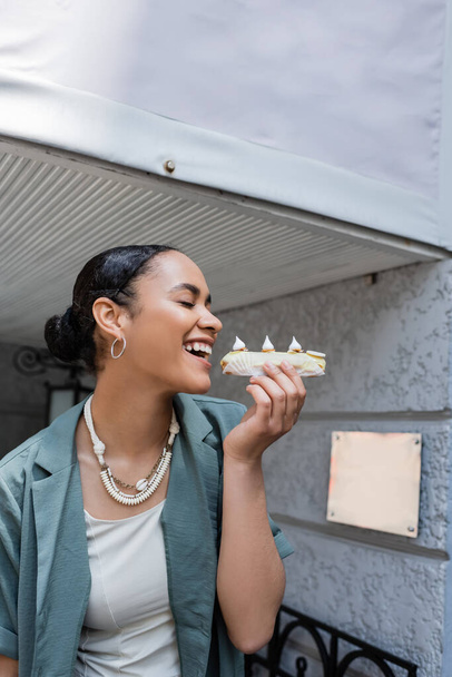 Smiling african american woman closing eyes while holding eclair near sweet shop outdoors  - Fotó, kép