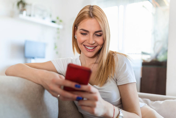 自宅のソファに座りながらスマートフォンを使った魅力的な笑顔の女性。コミュニケーションと居心地の概念. - 写真・画像