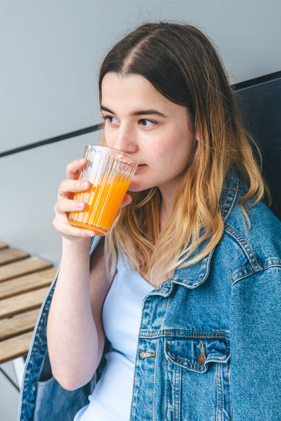A young woman in a blue denim jacket is drinking orange juice. - 写真・画像