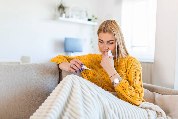 Sick desperate woman has coronavirus. Covid 19 , cold, sickness, allergy concept. Pretty sick woman has runnning nose, rubs nose with handkerchief and looking at temperature on thermometer - Fotografie, Obrázek