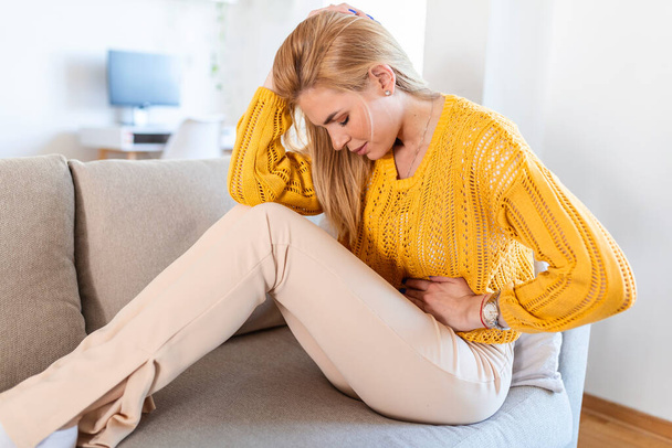 Young sick woman with hands holding pressing her crotch lower abdomen. Medical or gynecological problems, healthcare concept. Young woman suffering from abdominal pain while sitting on sofa at home - Foto, afbeelding