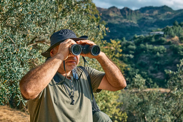 Tourist man looking through binoculars at the mountains view. Traveling with backpack or exercising outdoors. Active people, hiking, healthy lifestyle and harmony concept. Unity with nature. - Photo, Image