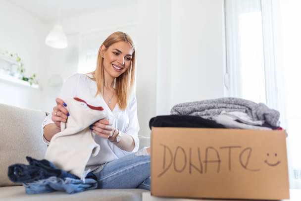 Young woman with donation box at home. Donation box for poor with clothing in female hands. Woman donates clothing to shelter. Cheerful woman holds box of donated clothing - Photo, Image