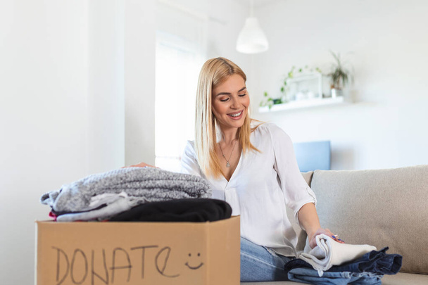 Glückliche junge Frau sitzt zu Hause auf Couch, steckt Kleidung in Spendenbox, kümmert sich um Frauen, steckt Kleidung in Kartons, spendet für Bedürftige, wiederverwendet, recycelt Konzept - Foto, Bild