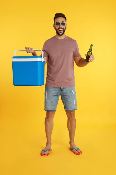 Happy man with cool box and bottle of beer on yellow background - Foto, imagen