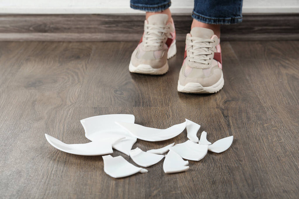 Woman in sneakers standing near broken plate on floor indoors, closeup - Zdjęcie, obraz