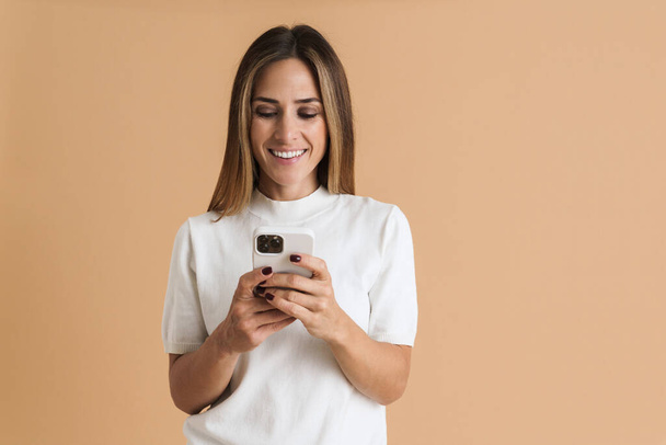 White mid woman wearing t-shirt smiling and using cellphone isolated over beige background - 写真・画像