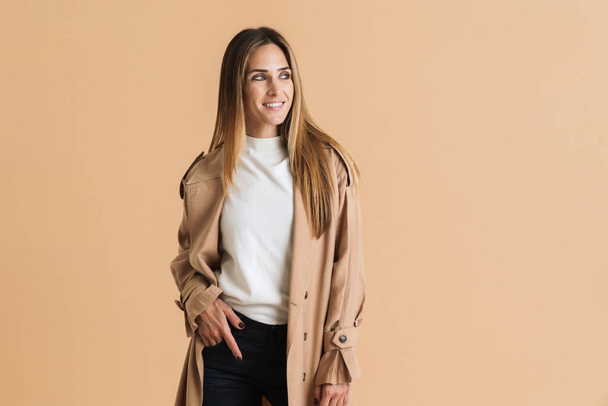 White mid woman wearing coat smiling and looking aside isolated over beige background - Foto, imagen