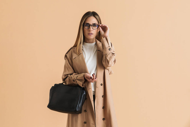 White mid woman wearing coat looking at camera while posing with bag isolated over beige background - Fotografie, Obrázek