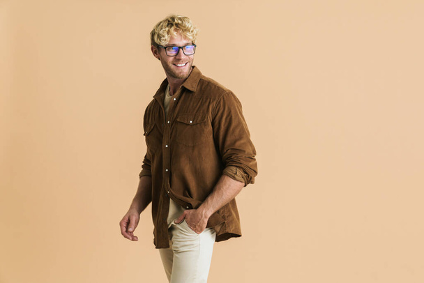 White blonde man in eyeglasses smiling and looking aside isolated over beige background - 写真・画像