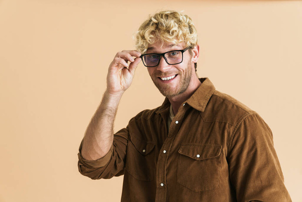 White blonde man in eyeglasses looking and smiling at camera isolated over beige background - Fotó, kép
