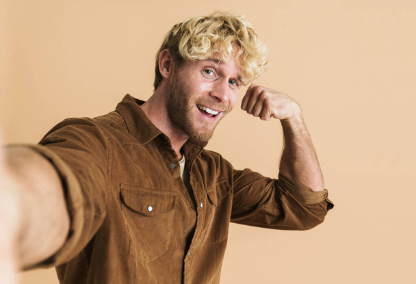 White blonde man showing his bicep while taking selfie photo isolated over beige background - Фото, зображення