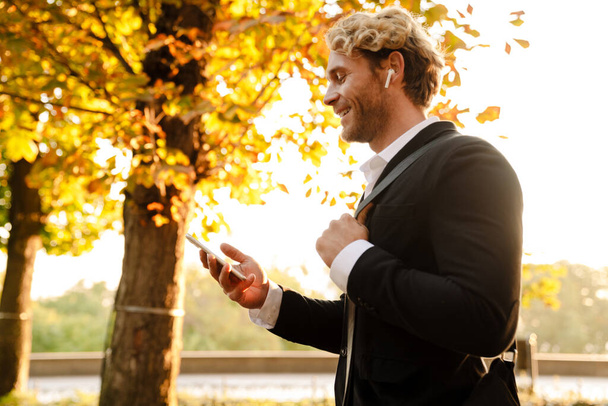 Blonde man using earphones and mobile phone while walking in autumn park - Foto, Bild