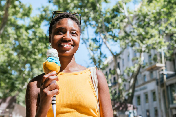 glimlachende Afrikaanse vrouw die op straat loopt met een ijsje op haar hand - Foto, afbeelding