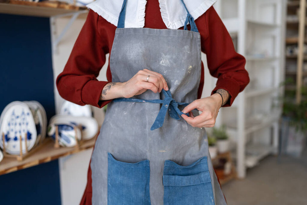 Female ceramist tying her apron before working with raw clay in pottery studio. Young student girl or master posing art dirty apron at her studio. Pottery business and workshop concept - Fotografie, Obrázek