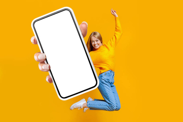 Fröhliche hübsche junge Dame springt in die Luft auf orangefarbenem Hintergrund mit Kopierraum, zeigt Handy mit weißem Leerbildschirm, genießt neueste mobile Anwendung, Attrappe, Collage - Foto, Bild