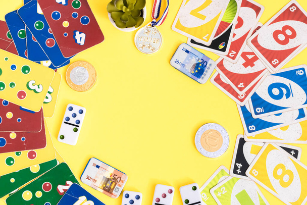 Old,dirty,colorful game cards ligretto uno,dominoes,chocolate money and winner medal lie in a round frame on a yellow background with copy space in the center,flat lay closeup.Summer board games concept. - Φωτογραφία, εικόνα