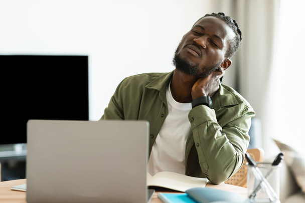 Neck ache. Upset african american guy massaging aching neck, suffering from pain after computer work, sitting workplace at home office. Poor posture issue and arthritis problem - 写真・画像