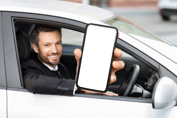 Mobile app for car rental, car parking. Cheerful businessman sitting in luxury white car, showing cell phone with white blank screen, through auto window shot, copy space, mockup, collage - Fotoğraf, Görsel