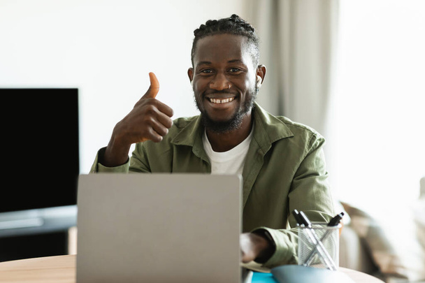 Szczęśliwy czarny mężczyzna freelancer pracujący na laptopie i gestykulujący kciuki w górę, polecający pracę na odległość, uśmiechający się do kamery w domu. Lubię zdalną pracę. - Zdjęcie, obraz