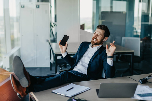 Overjoyed businessman relaxing in office, resting feet on table and enjoying music in earphones, holding cellphone and dancing, taking break from work, copy space - Foto, immagini