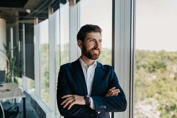Portrait of handsome caucasian businessman standing with folded arms near window, successful entrepreneur having break in office, advertising professional services, free space - Foto, Bild