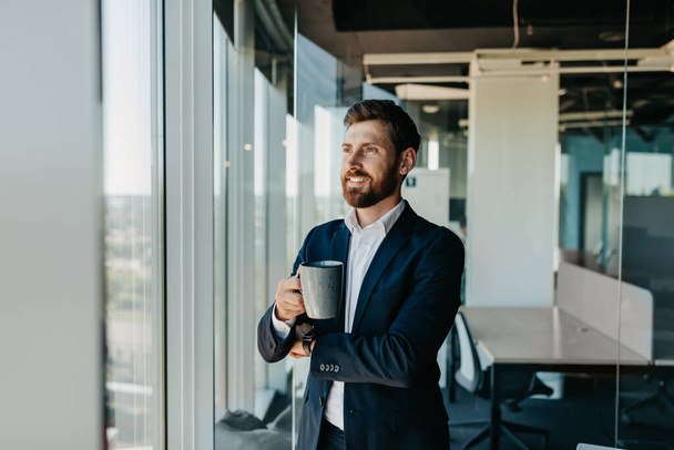 Красивый успешный бизнесмен с кофе-брейком, стоя рядом с окном в своем офисе с чашкой кофе, глядя в сторону и улыбаясь, бесплатное пространство для копирования - Фото, изображение