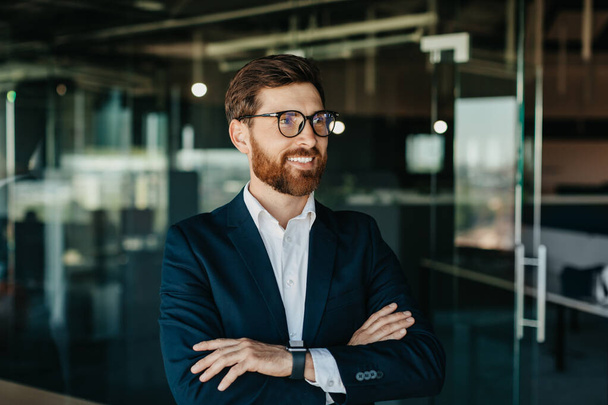 Porträt eines erfolgreichen Geschäftsmannes im Anzug, der mit verschränkten Händen posiert, wegschaut und lächelt, im modernen Bürointerieur arbeitet, freier Kopierraum - Foto, Bild