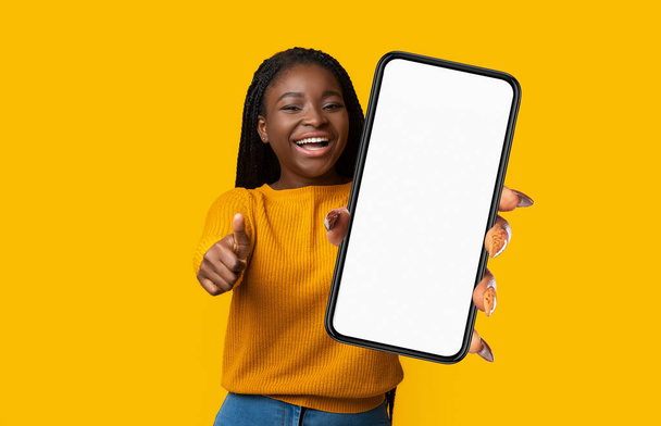 Positive Young Black Woman Showing Smartphone With Empty Screen And Gesturing Thumb Up At Camera, Posing Over Yellow Studio Background, Beautiful Young Female Having Fun, Copy Space, Mockup, Collage - Фото, изображение