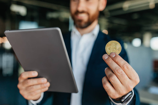 Confident businessman holding golden bitcoin and working with charts on digital tablet, standing in office, crop, selective focus. Bitcoin cryptocurrency investment concept - Foto, Bild