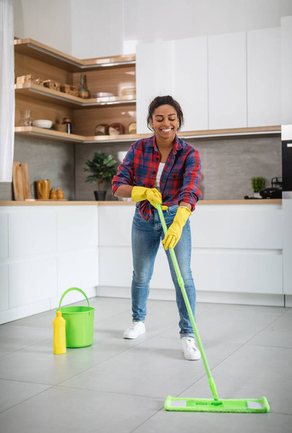 Iloinen tuhatvuotinen afrikkalainen amerikkalainen kotiäiti kumihanskat mopilla pesee lattian viihtyisä keittiö sisustus, pystysuora, vapaa tila. Hygienia ja kotityöt. Aktiivinen innoissaan taloudenhoitaja do kotitalous - Valokuva, kuva