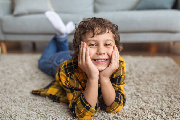 Szczęśliwego dzieciństwa. Zbliżenie portret wesołego chłopca uśmiechniętego do kamery, leżącego na podłodze w domu, opierającego się o podbródek na rękach - Zdjęcie, obraz