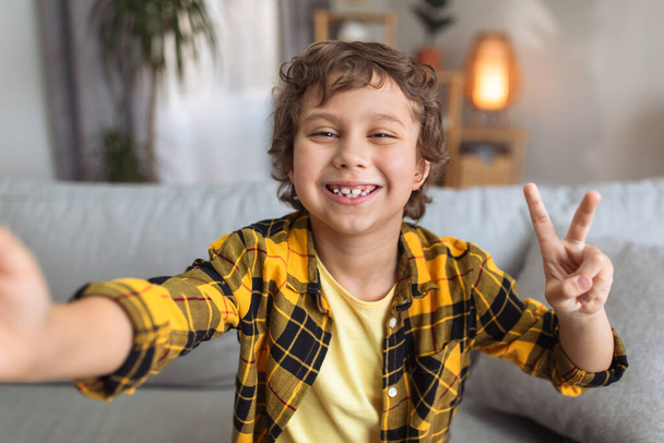 Selfie time. Camera pov portrait of happy little boy blogger making photo, smiling to camera and showing peace gesture, having fun at home - Фото, изображение