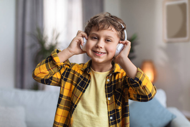 Nahaufnahme Porträt eines entzückenden glücklichen kleinen Jungen, der Musik über drahtlose Kopfhörer genießt, lächelnd in die Kamera lächelt und zu Hause im Inneren posiert - Foto, Bild