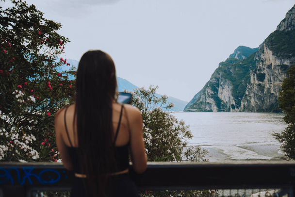A woman takes a photo on the shores of Riva Del Garda lake and mountains in the background. Tourist in Italy. beauty of nature - 写真・画像