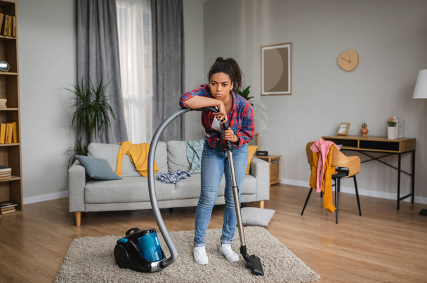 Unhappy tired sad millennial african american housewife vacuuming and rest from cleaning in living room interior. Household chores, troubles in domestic work, disorder at home, chaos with clothes - Zdjęcie, obraz