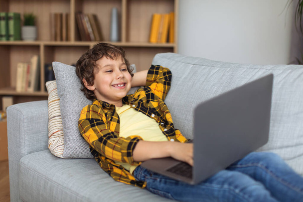 Leisure of modern kids. Happy little boy watching streaming video online on laptop, resting on sofa at home and smiling, free space - Foto, Imagen