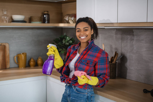 Glad pretty millennial african american housewife in rubber gloves and shirt with spray and rag in cozy kitchen interior, free space. House cleaning service, domestic work, purity and household chores - Photo, Image
