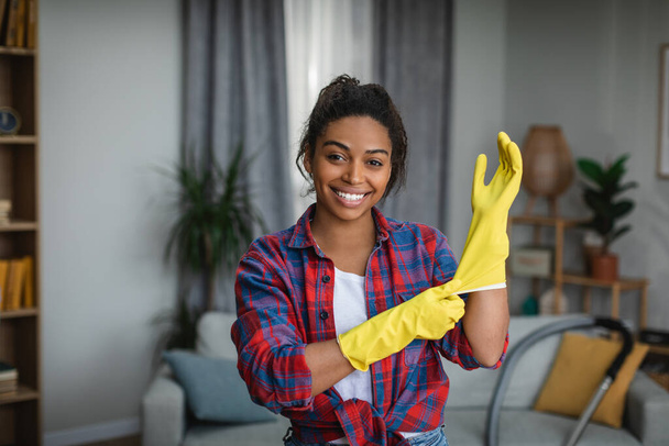 Smiling young african american woman puts on rubber gloves and ready to homework in living room interior. Housewife has fun and many household chores, domestic work and professional cleaning service - Foto, immagini