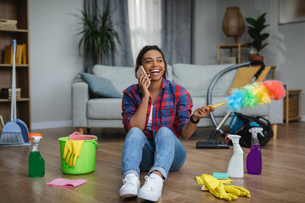 Fröhliche afrikanisch-amerikanische Hausfrau ruft per Smartphone an und sitzt mit Putzutensilien auf dem Boden im Wohnzimmer. Kommunikation bei Hausarbeit, Hausarbeit, Reinigung und Gadget - Foto, Bild
