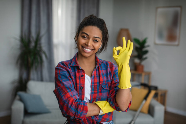 Glad millennial black lady in rubber gloves shows ok sign with hand in living room interior. Housewife has many household chores, house cleaning and excellent result professional cleaning service - Foto, Imagen