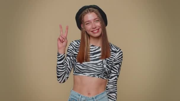 Lovely happy young woman showing victory sign, hoping for success and win, doing peace gesture, smiling with kind optimistic expression. Adult stylish girl isolated on beige studio background indoors - Záběry, video