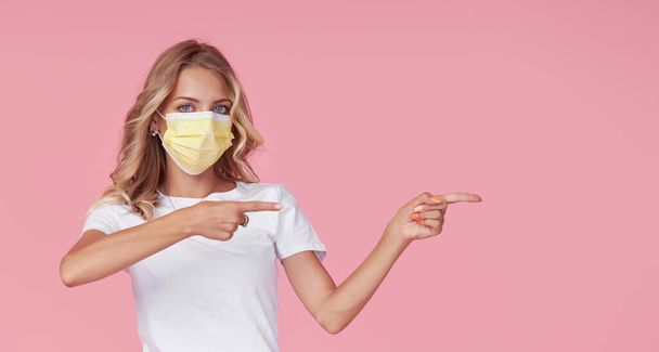 A young attractive woman in a white casual t-shirt wears a protective face mask. Protection against flu and cold diseases during a pandemic. Pointing to the right. Pink isolated background - Photo, Image
