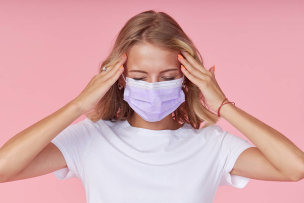 Young ill woman in a white casual t-shirt wears a protective face mask. holding hands on head. Protection against flu and cold diseases during a pandemic. Pink isolated background - Fotografie, Obrázek