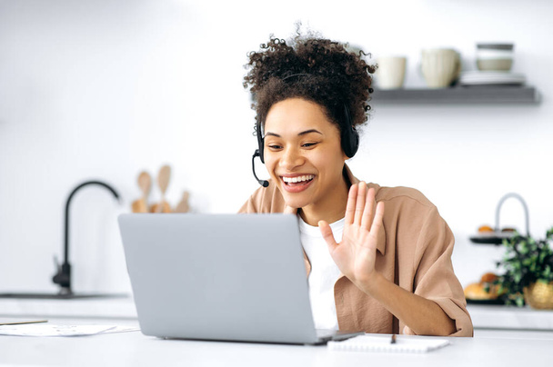 Mutlu Afro-Amerikan kıvırcık saçlı genç kadın, kulaklık ve dizüstü bilgisayar kullanıyor, bir video konferansında müşteriyle veya meslektaşıyla konuşuyor, mutfakta oturuyor, arkadaşça gülümsüyor. - Fotoğraf, Görsel