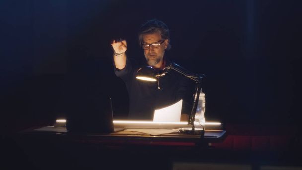 Director in glasses reading the script of the play under the lamp and looking at the camera, while sitting at the table in the dark hall of the theater - Fotografie, Obrázek
