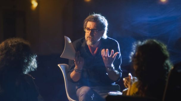 Mature director on a dim theater stage talking to the actor and disabled actress about the performance script, and showing how to act more emotionally - Valokuva, kuva
