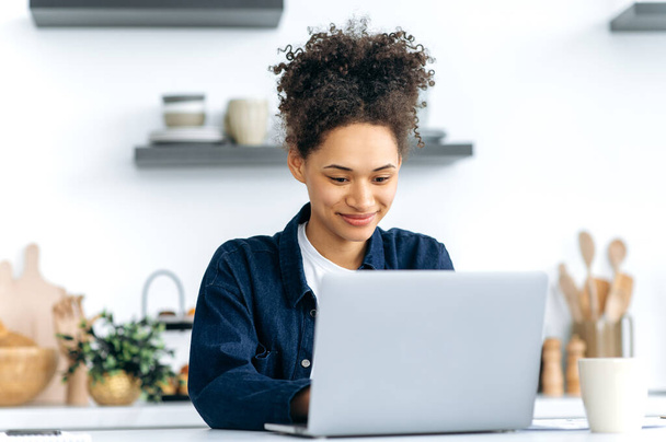 Muito bem sucedida jovem afro-americana, freelancer, especialista em TI, sentado em casa na cozinha, usando laptop para o trabalho, mensagens de texto com colegas, trabalhando em um projeto, sorrindo alegremente - Foto, Imagem