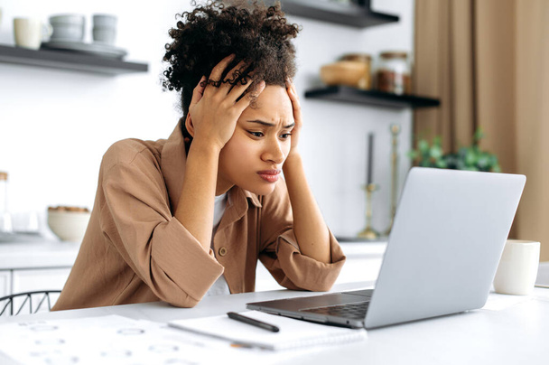 Desperate worried sad african american young woman, using laptop for work or study from home, sitting in kitchen, frustrated looking at screen, failed project, got financial loss, job downgrade - Fotó, kép