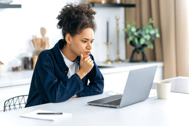 An anxious African American woman, trader, freelancer, looks worriedly at the laptop screen, monitors the situation in the financial market, concentrated analyzes the dynamics, feeling stressful - Photo, image